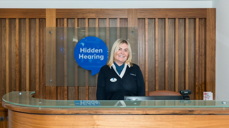 Interior photograph of the Hidden Hearing newbridge reception with clinic coordinator Sharon standing in front of Hidden Hearing signage