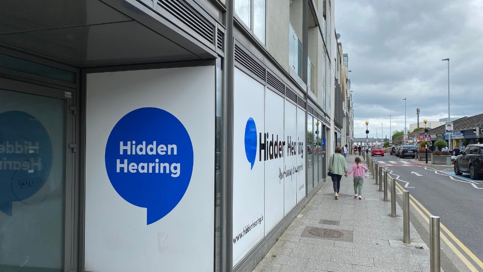 The storefront of Hidden Hearing, a hearing clinic, features a large blue speech bubble logo with the text 'Hidden Hearing.' A mother and child walk on the sidewalk alongside a city street lined with buildings and parked cars. The website www.hiddenhearing.ie is visible on the display.