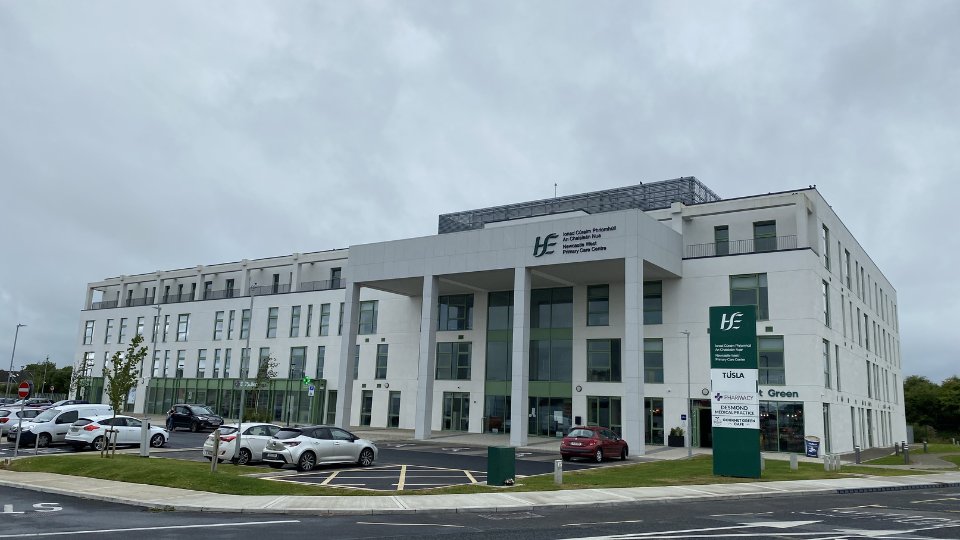 A modern white healthcare building labeled 'HSE,' featuring large glass windows, surrounded by parked cars and greenery. Sign reads: 'VCarl Corbitt Residential / Adult Mental Health Unit / Ennis Primary Care Centre,' along with other service listings including 'Tusla,' 'Pharmacy,' and more.