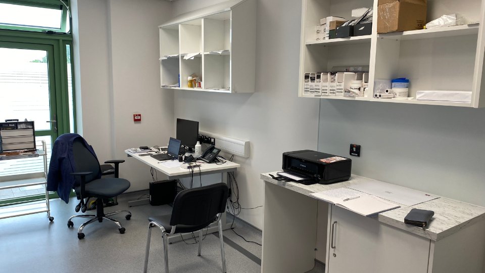A modern hearing clinic room with a desk featuring a computer and equipment, shelves holding hearing care supplies, a printer on the countertop, and a green-framed window illuminating the space. No text present.