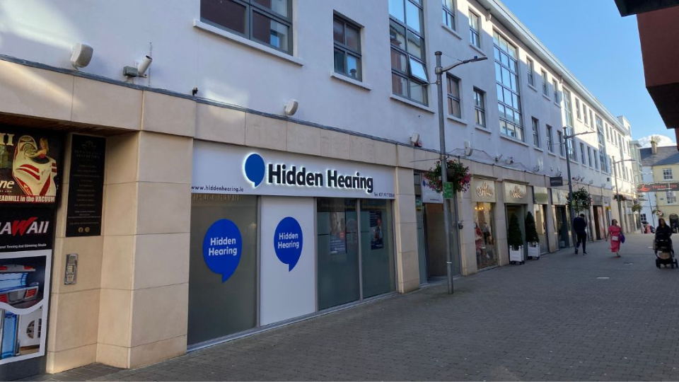 Hidden Hearing storefront displaying the brand name in bold blue and black text, with 'Hidden Hearing' logos on the glass windows, set in a pedestrian shopping street with nearby businesses. Website: www.hiddenhearing.ie.