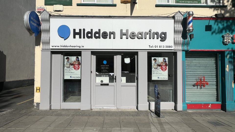 The storefront of Hidden Hearing, a hearing clinic, features a white sign with a blue logo and black text reading 'Hidden Hearing' and 'Tel: 01 813 5880.' Posters on the windows advertise free hearing tests. The website www.hiddenhearing.ie is also displayed prominently. Grey double doors sit between large windows, with a pavement sign in front. The building is flanked by a barber shop and other businesses.