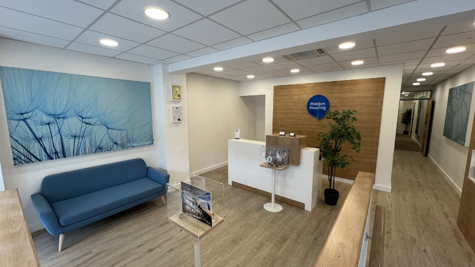 A modern hearing clinic reception area features a blue sofa, wood accents, and a 'Hidden Hearing' branded sign. Brochures and displays about hearing aids stand near the front desk.