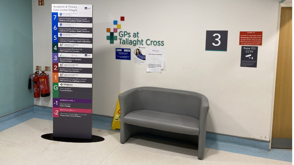 A directory pillar listing services at the Academic & Primary Care Centre Tallaght stands near a gray cushioned bench. The 'GPs at Tallaght Cross' logo is on the wall, alongside COVID-19 signage and directional panels.
