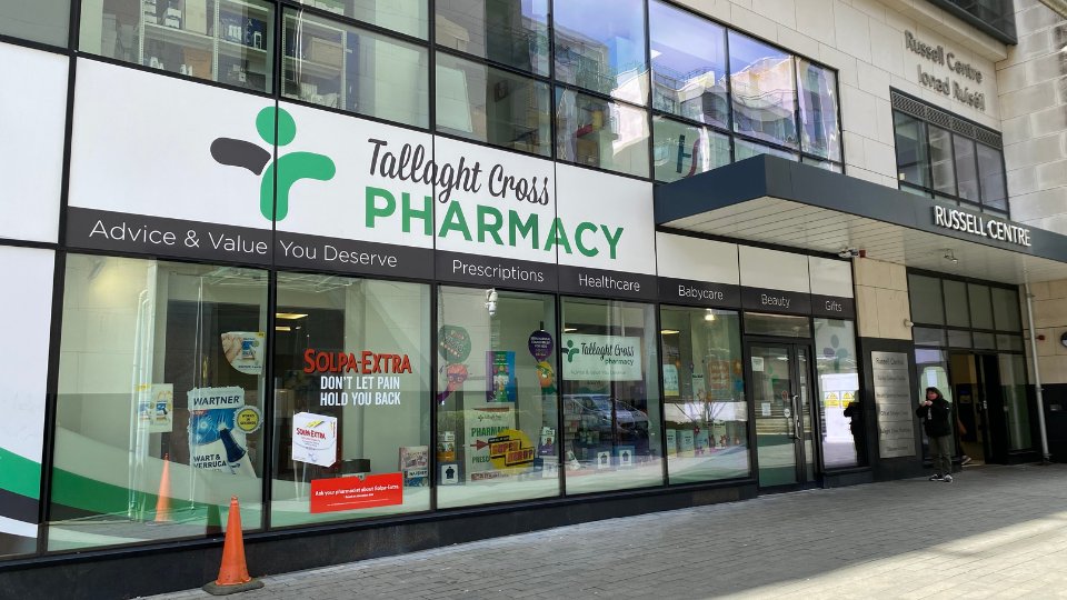 Tallaght Cross Pharmacy storefront displays a clean aesthetic, branded with green and white signage, promoting services like prescriptions, healthcare, and beauty products. Large windows showcase product displays. Text includes:  
                        
                        - 'Tallaght Cross PHARMACY'  
                        - 'Advice & Value You Deserve'  
                        - 'Prescriptions, Healthcare, Babycare, Beauty, Gifts'  
                        - 'SOLPA-EXTRA DON'T LET PAIN HOLD YOU BACK'  
                        
                        The pharmacy is located within the 'Russell Centre,' as labeled on the building. A pedestrian walks near the entrance.