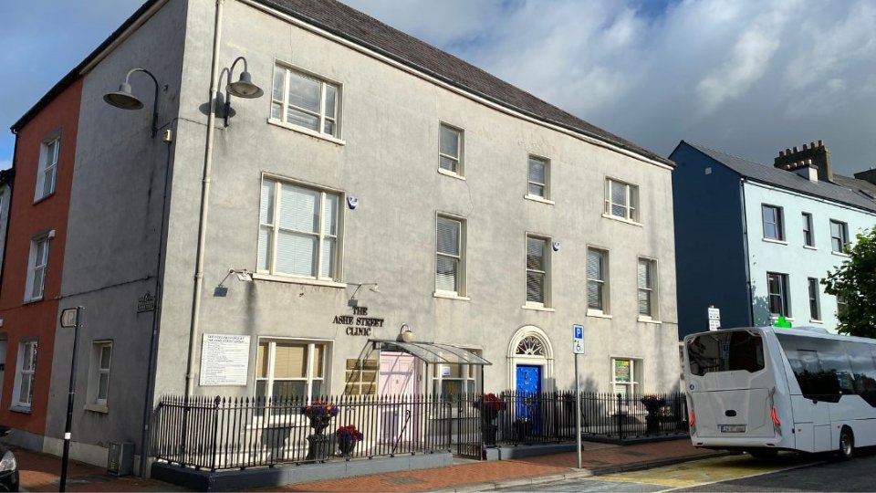 A three-story beige building labeled 'The Ashe Street Clinic' with a blue door, surrounded by a black fence, adjacent colorful buildings, a parked white bus, and a cloudy sky. Signs and plaques are visible but not legible.