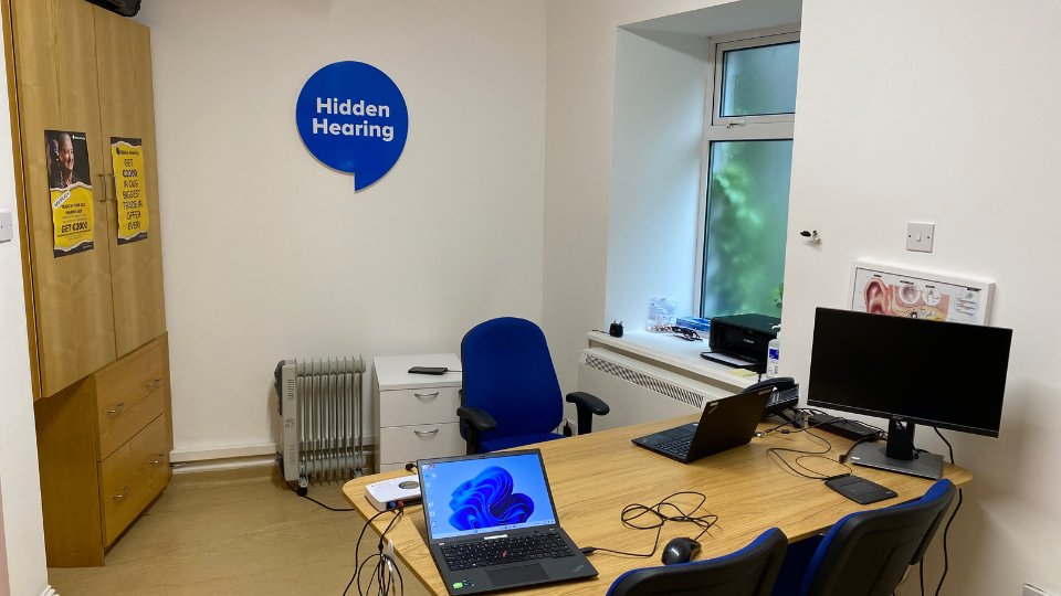 A Hidden Hearing consultation room featuring a desk with two laptops, a computer monitor, blue chairs, wall posters about ear health, cabinets, and a visible 'Hidden Hearing' logo on the wall.