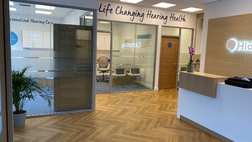 [GEN] A welcoming hearing clinic features a reception desk with a 'Hidden Hearing' illuminated logo, glass-walled consultation rooms labeled 'Life Changing Hearing Health,' hardwood floors, and comfortable seating. Text includes: 'Personalised Hearing Care' and 'We create trust.'