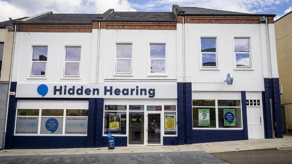 [GEN] The Hidden Hearing clinic storefront features a white façade with navy blue accents, displaying signage for 'Hidden Hearing' and offering 'FREE Hearing Tests' and 'Personalised Hearing Care.' Contact: Tel: 051 378 877, www.hiddenhearing.ie.