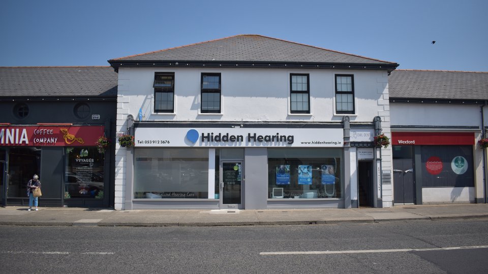[GEN] The storefront for Hidden Hearing features a clean, white façade with the company name, logo, and text: 'Tel: 053 912 3676' and 'www.hiddenhearing.ie.' Located between a coffee shop and another business, it displays hearing care advertisements in large front windows, promoting professional hearing aid solutions and services in Wexford.