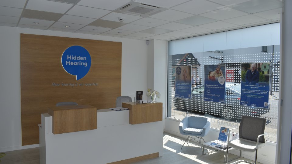 A modern Hidden Hearing clinic reception features a wooden panel with the logo 'Hidden Hearing' and tagline 'Your hearing is our expertise.' The space includes a white desk, decorative flowers, and a seating area with chairs and brochures. Large windows display promotional posters addressing hearing aids, hearing tests, and hearing care services, with street views visible outside.