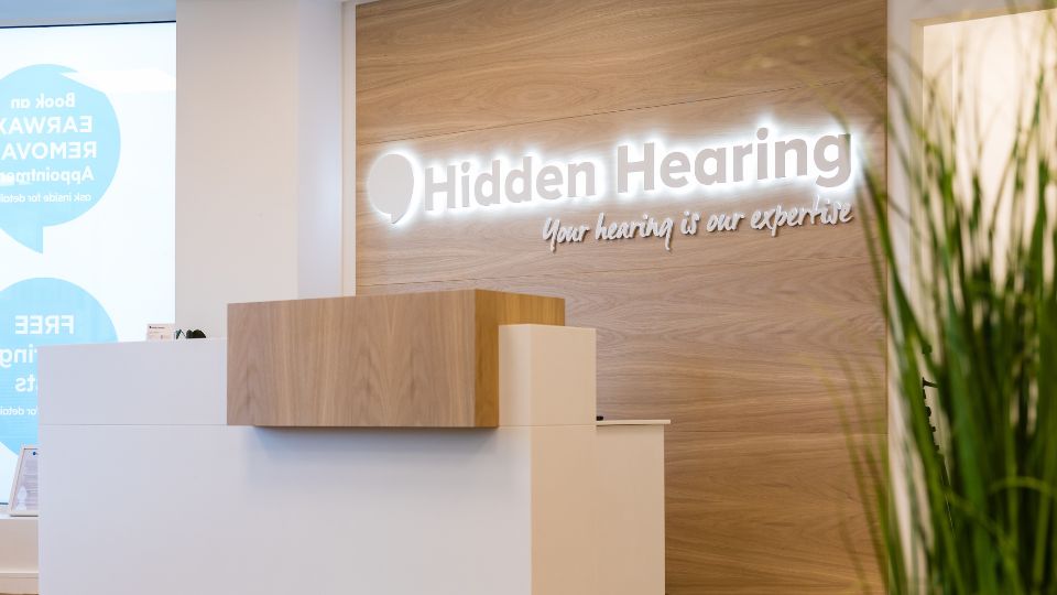 Interior photograph of a Hidden Hearing clinic reception with a illuminated Hidden Hearing sign behind the counter