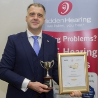 A suited individual holds a trophy and a framed certificate, standing before a Hidden Hearing-branded banner displaying the tagline 'We listen, you hear' and text referencing hearing problems and clinics.