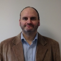 A person wearing a brown blazer over a light blue collared shirt stands against a plain, light-colored wall. The face is blurred for privacy.