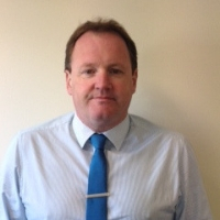 A person wearing a light-striped collared shirt and a blue tie with a tie bar stands against a plain beige background. No text or hearing aid brands are visible in the image.