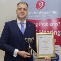 A suited individual holds a trophy and a framed certificate, standing before a Hidden Hearing-branded banner displaying the tagline 'We listen, you hear' and text referencing hearing problems and clinics.