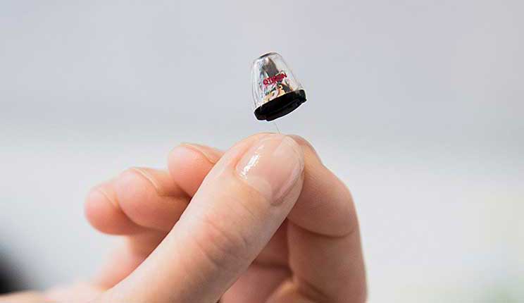 [GEN] A small, in-the-ear hearing aid held between two fingers; the transparent casing reveals inner components. The background is softly blurred, suggesting a clinical setting like a hearing clinic. No text is visible in the image.