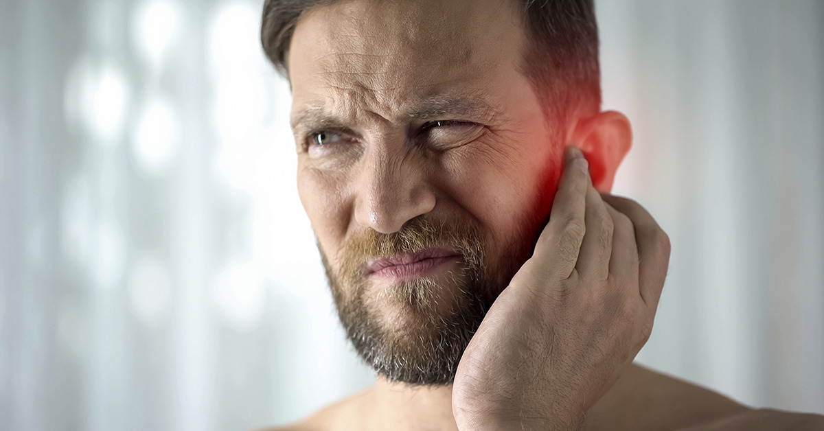 A man holds his hand to his ear, which appears red, possibly indicating discomfort or hearing issues, against a softly blurred background.