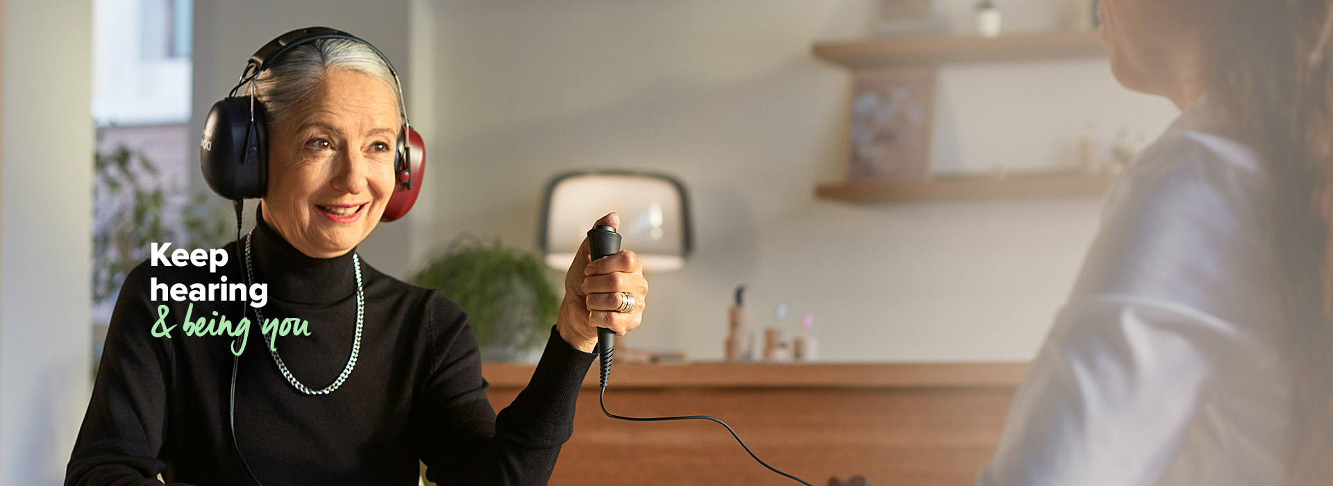 A person undergoing a hearing test wears headphones while holding a response button, seated in a modern hearing clinic. Text reads: 'Keep hearing & being you.'
