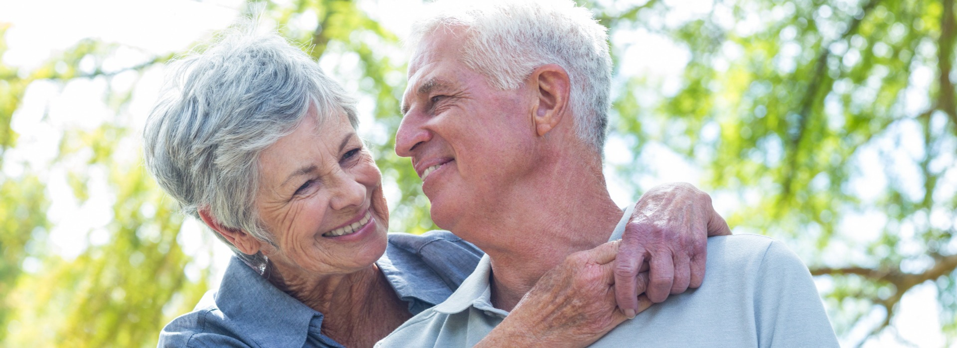 An elderly couple stands closely outdoors under sunlight, with blurred faces surrounded by lush green foliage. The man’s ear, central in the frame, suggests themes like hearing care or hearing aids.