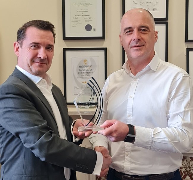 Two individuals shake hands as one presents a glass award in an office, with framed certificates, including an 'Audiologist Certificate of Commendation,' displayed on the wall in the background.