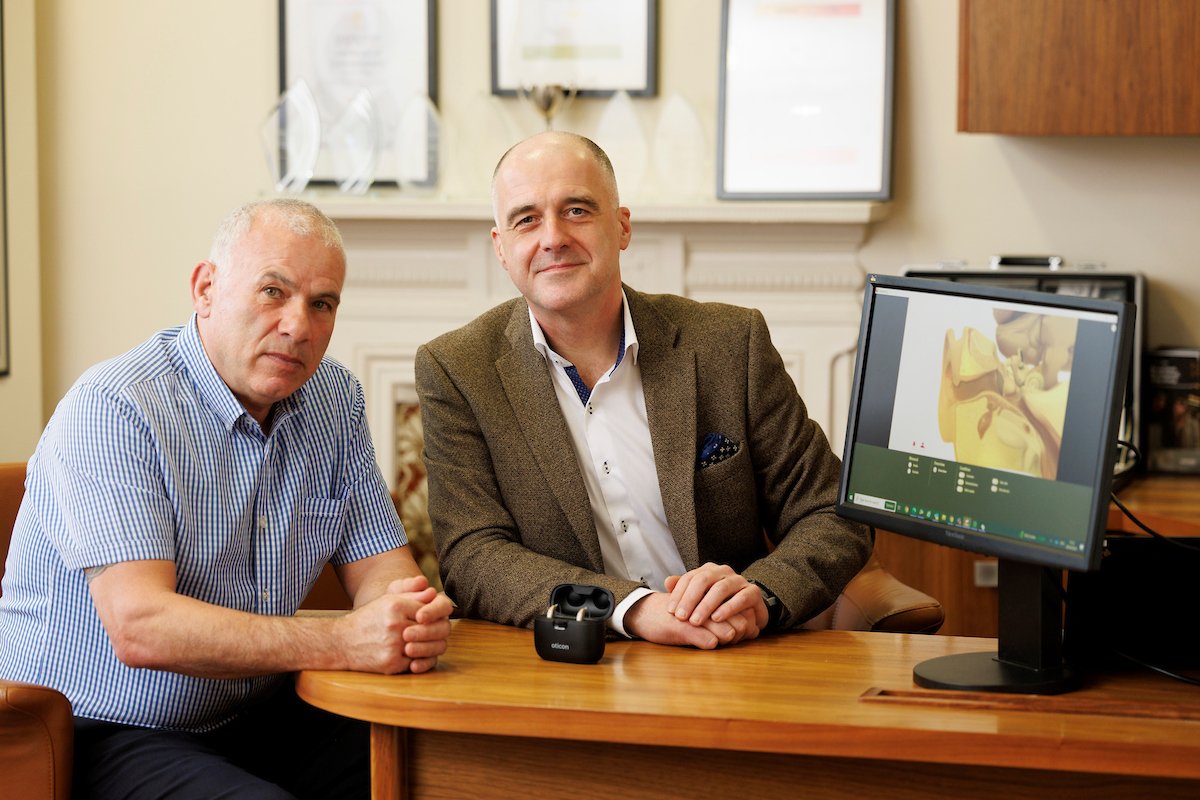 Two men sit at a wooden desk in a hearing clinic. A computer screen displays an ear anatomy diagram, while an Oticon hearing aid case rests on the desk. Framed certificates decorate the background.