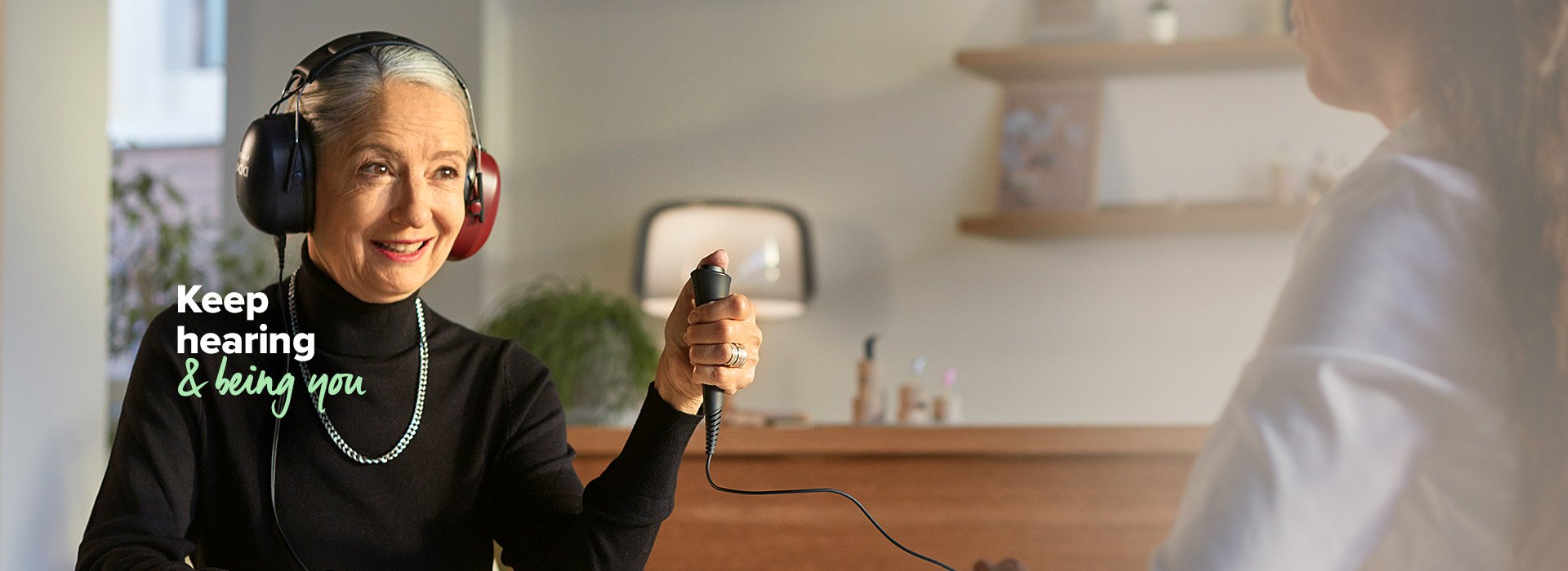 A person undergoing a hearing test wears headphones while holding a response button, seated in a modern hearing clinic. Text reads: 'Keep hearing & being you.'