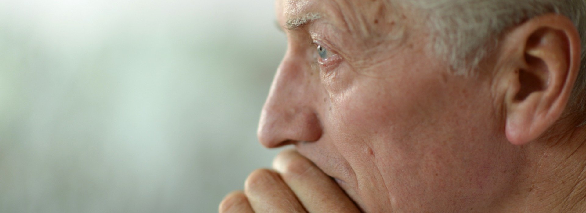 A close-up shot of an older person's right ear, surrounded by short gray hair, set against a softly blurred background of neutral green tones, suggesting a calm indoor environment.