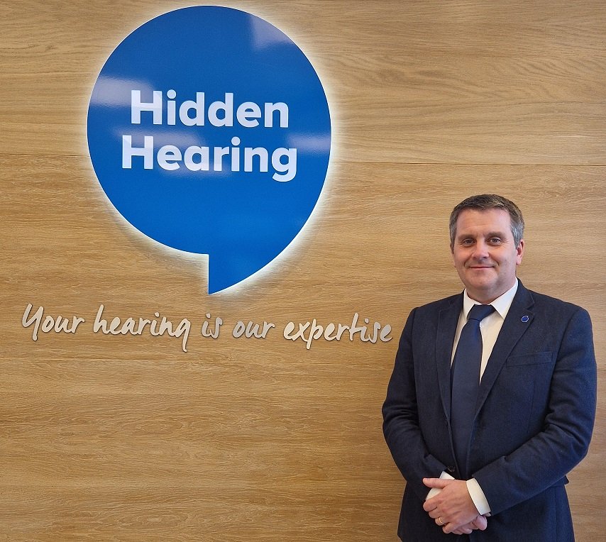A suited professional stands in front of a wooden backdrop featuring Hidden Hearing’s blue speech bubble logo and tagline, 'Your hearing is our expertise,' representing a hearing clinic specializing in hearing care.