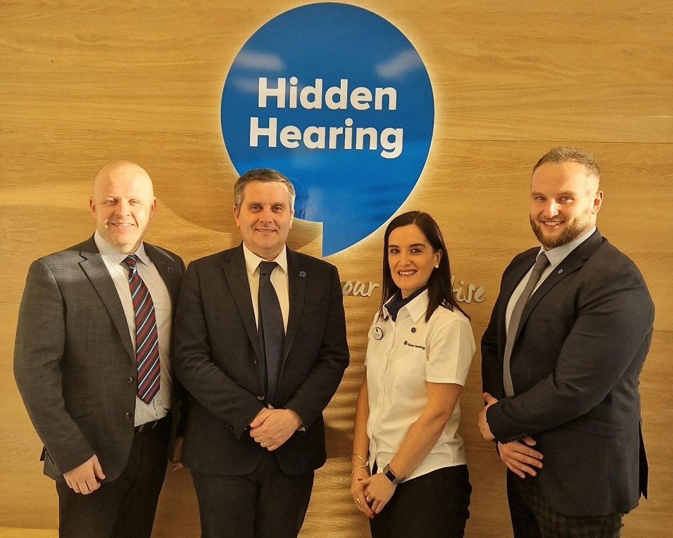 Four individuals in formal attire stand in front of a wooden wall with a large blue balloon-shaped logo reading 'Hidden Hearing' and the tagline 'Life is worth hearing.' The setting is a professional hearing clinic.