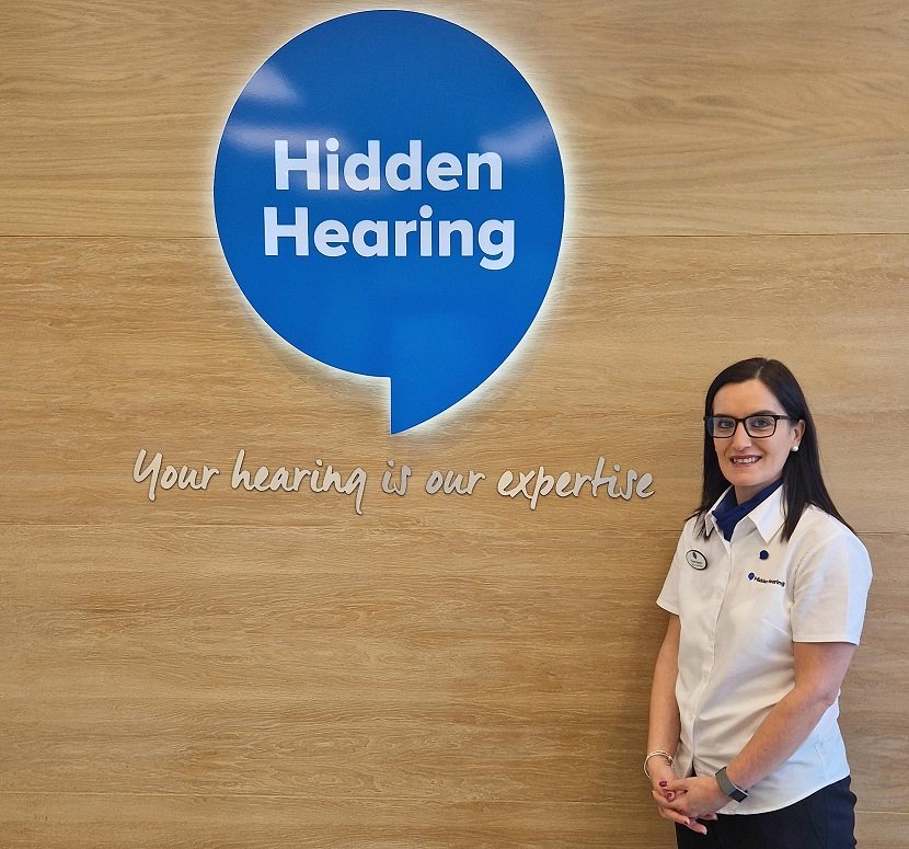 A Hidden Hearing staff member, wearing a branded uniform, stands beside a large blue sign with the text 'Hidden Hearing' and the slogan 'Your hearing is our expertise,' against a wooden wall.