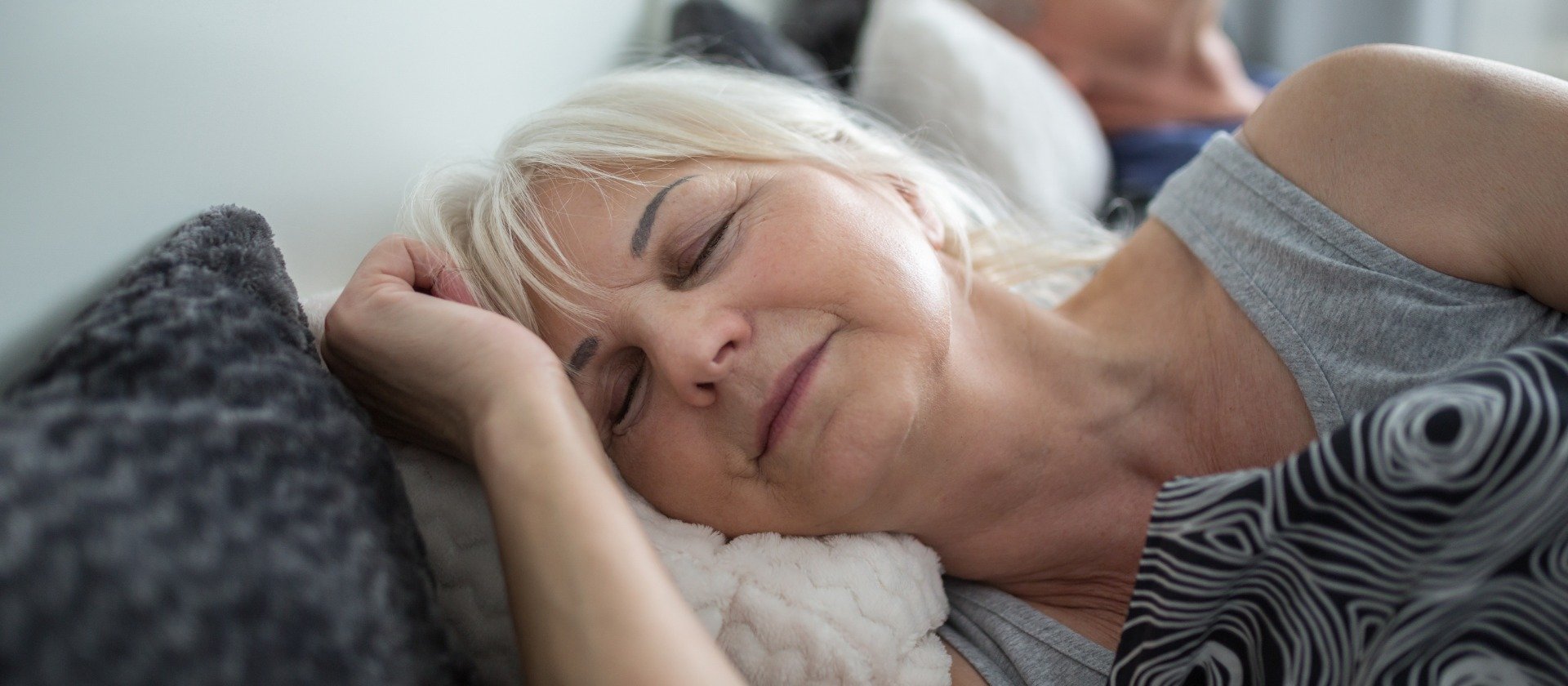 A person with light gray hair lies sideways on a bed, resting on patterned pillows and covered with soft blankets, in a cozy, indoor setting.