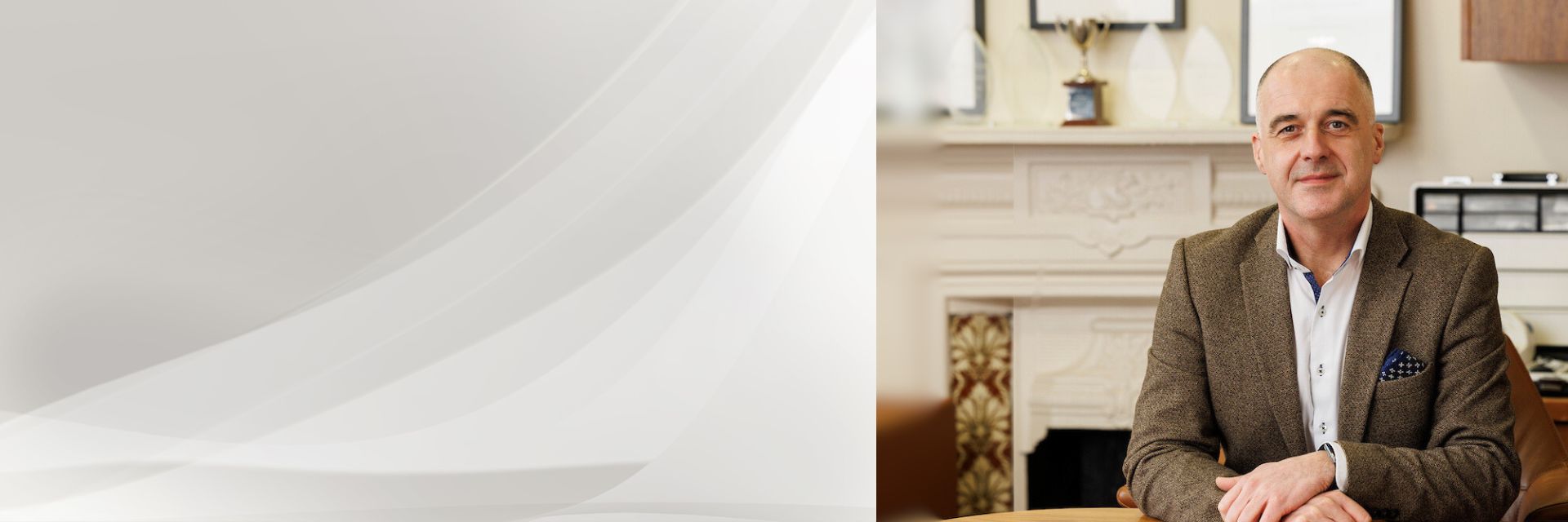 A man in a brown blazer sits at a wooden desk with a blurred face; the background features a fireplace, framed certificates, trophies, and office decor, suggesting a professional setting.