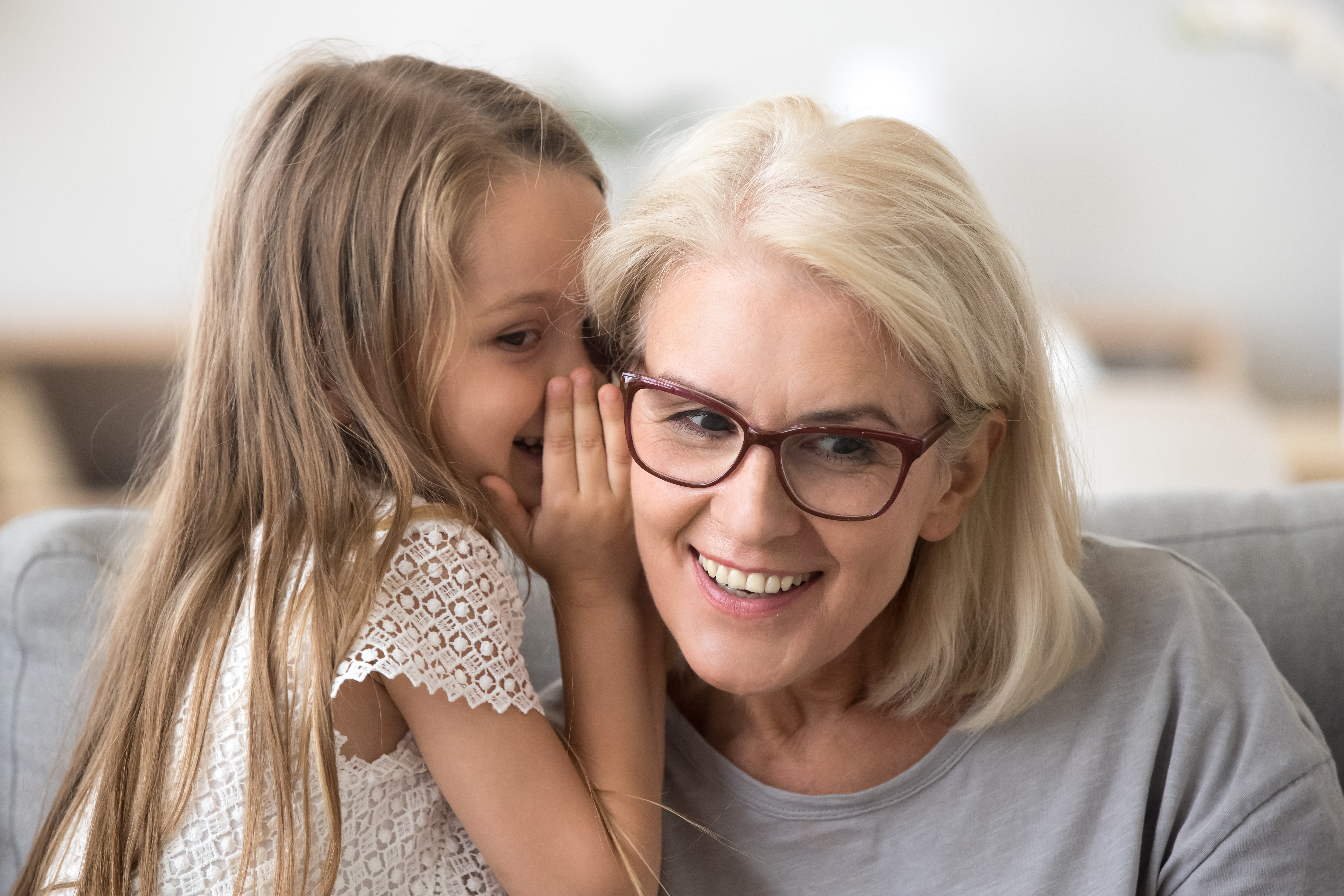 A young girl whispers closely to an older woman with gray hair on a sofa in a bright home setting, fostering an intimate moment of communication and connection.