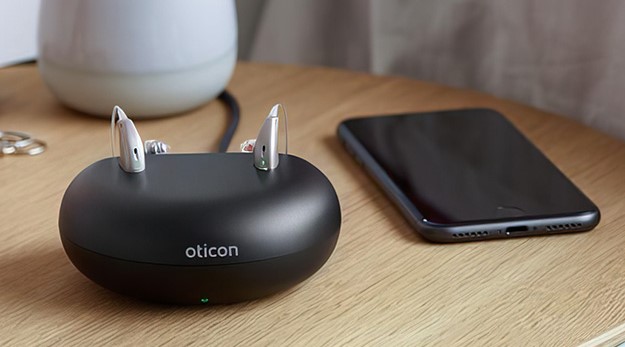 Rechargeable hearing aid charging station with Oticon logo placed beside a smartphone on a wooden surface.