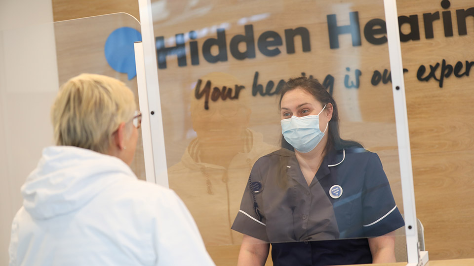 A professional audiology receptionist interacts with a client across a protective screen in a Hidden Hearing clinic, with the slogan 'Your hearing is our expertise' featured on a wooden background wall.