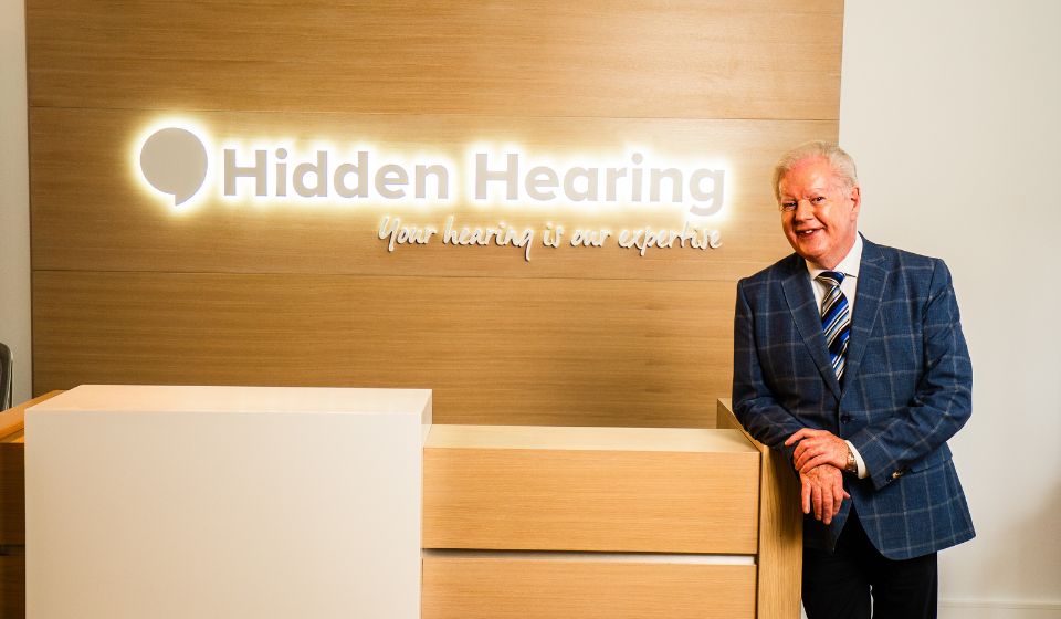 Broadcaster Julian Simmons in a suit smiling while leaning on a countertop with an illuminated Hidden Hearing sign behind him