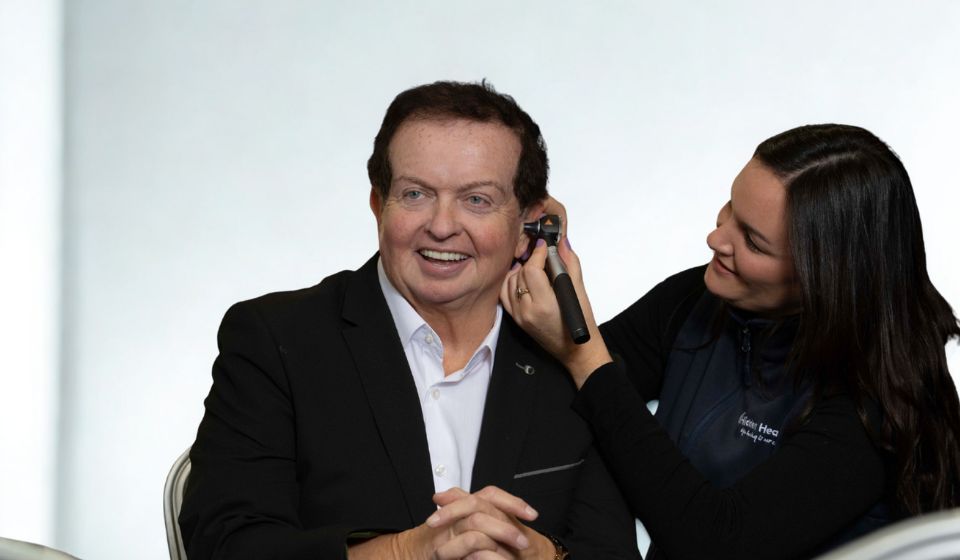 Marty Morrissey in a black suit getting his ear examined by an audiologist with an otoscope