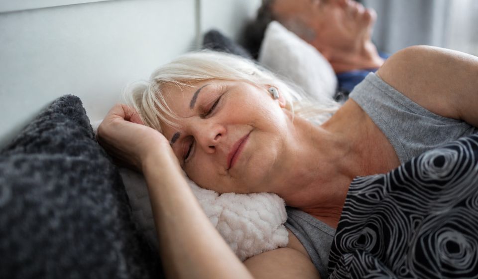 A woman sleeping peacefully in bed while listening to something on her headphones while a man sleeps behind her