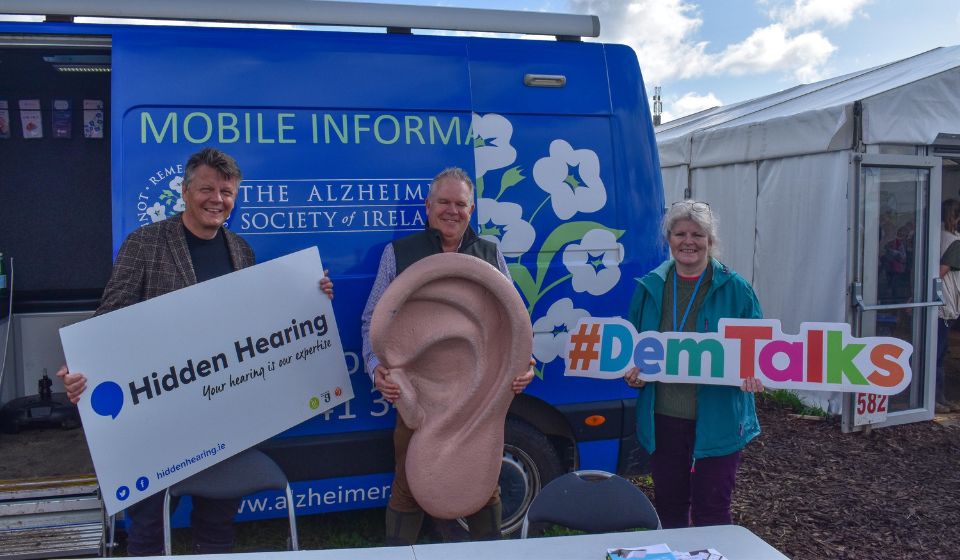 Three individuals hold props, including a large ear replica, a 'Hidden Hearing' sign, and a '#DemTalks' sign, in front of an Alzheimer Society of Ireland mobile information van beside a white event tent. Text on the Hidden Hearing sign reads:  
'Hidden Hearing'  
''Your hearing is our expertise''  
'hiddenhearing.ie'  