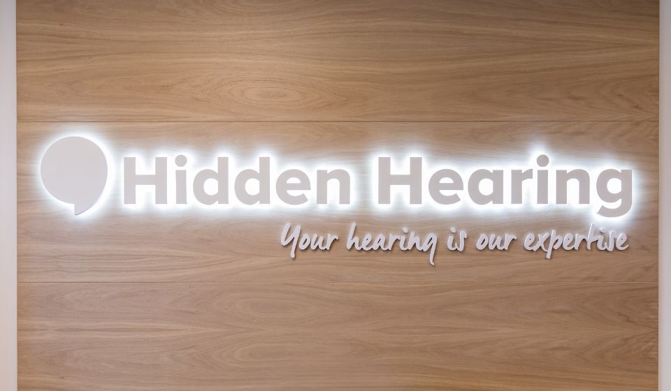 Hidden Hearing logo illuminated on a wooden wall with the tagline 'Your hearing is our expertise.'