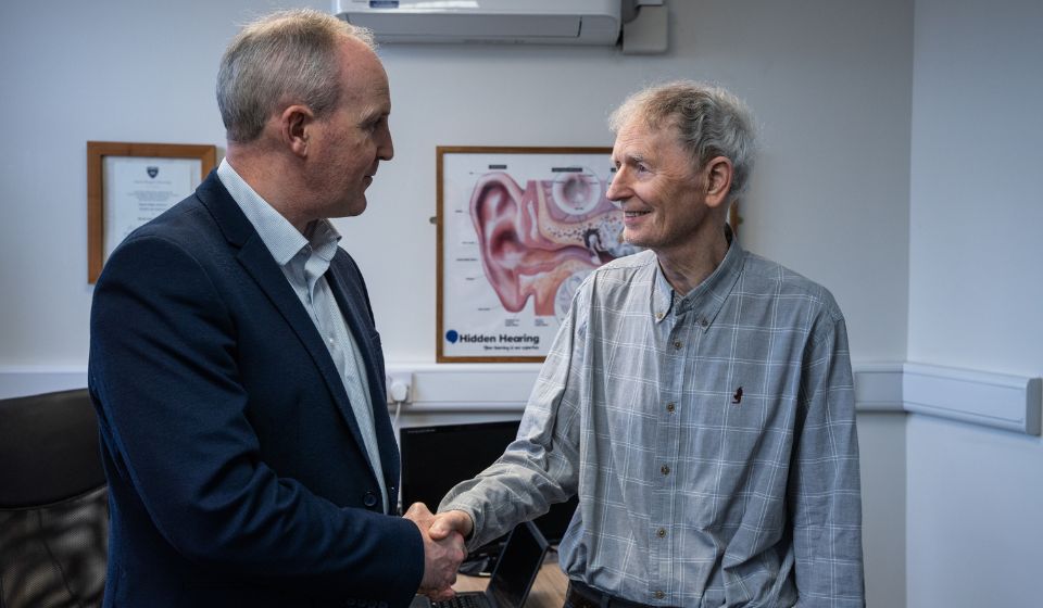 Audiologist shaking hands with patient in a consultation room with a anatomical graphic of an ear on the wall behind them