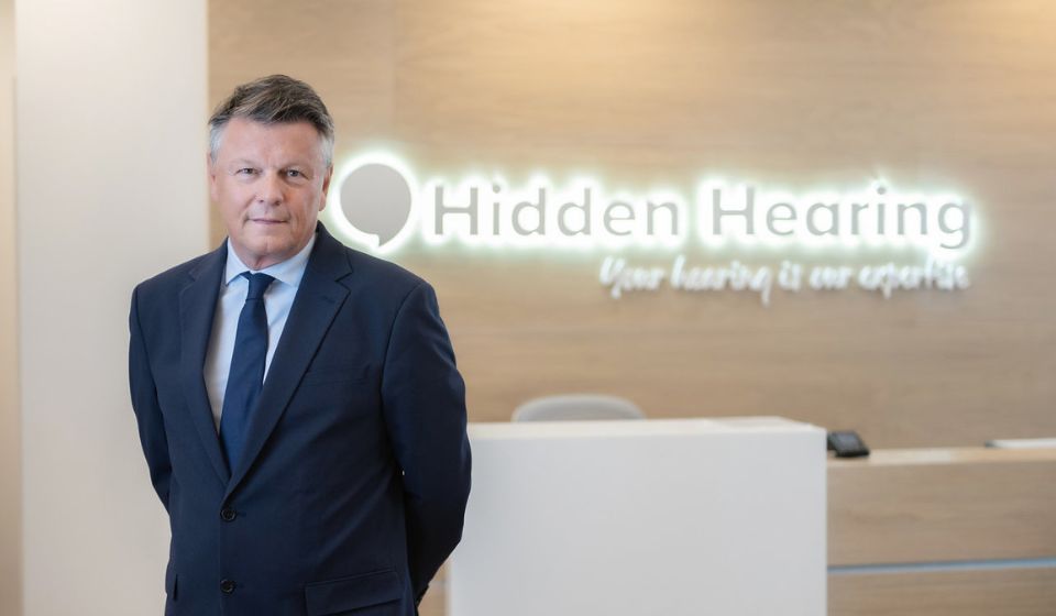 Managing Director Duncan Graham posing in a black suit in from of a bright Hidden Hearing sign