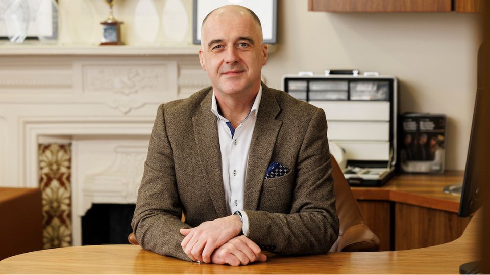 [GEN] A professionally dressed individual in a brown suit jacket sits at a wooden desk in a sophisticated office with a fireplace, trophy, and cabinetry. Hearing-related materials are visible on shelves.