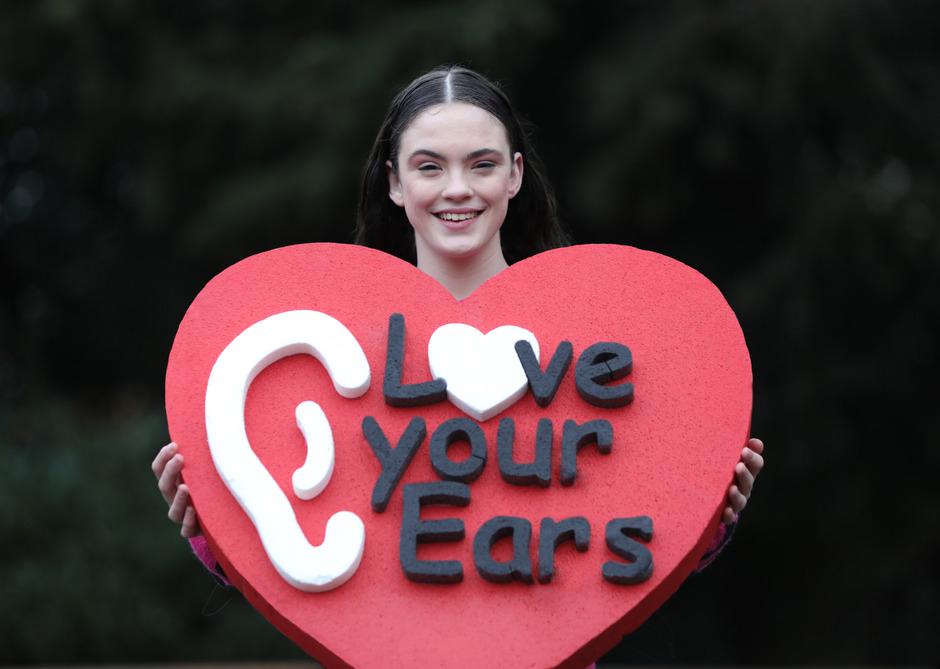 A person holds a large red heart-shaped sign featuring a white ear icon, a white heart, and the text 'Love your Ears,' set against an outdoor, blurred green backdrop.