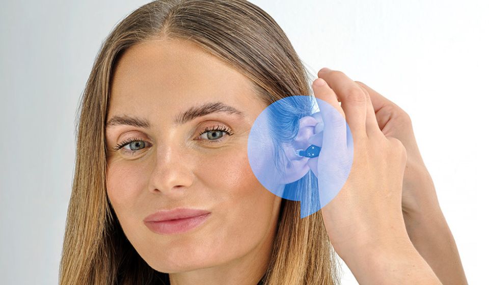 A photograph of a woman being fitted with the brand new nearly invisible Oticon Zeal hearing aid with a blue speech bubble over her ear