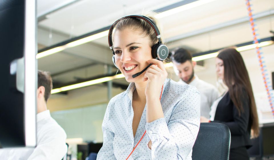 A professional wearing over-ear headphones interacts while seated in a modern office environment with bright lighting. Colleagues in business attire work in the background, creating a collaborative workspace.