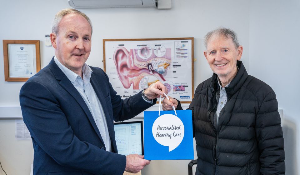 Audiologist David Courtney smiling while giving a bag labelled 'Personalised Hearing Care' to Tommy Swarbrigg in a Hidden Hearing consultation room with an ear anatomy poster on the wall