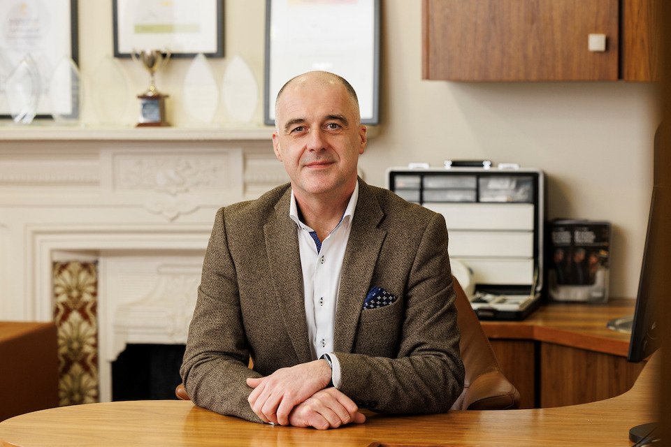 A professionally dressed individual sits at a wooden desk in a well-appointed office featuring framed certificates, a trophy, and hearing care materials. The scene likely depicts a hearing clinic consultation.