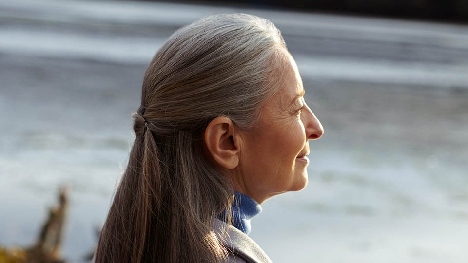 A woman with long, graying hair tied back gazes toward a calm, waterfront setting. Her visible ear suggests the potential use of a discreet hearing aid, emphasizing hearing care solutions.